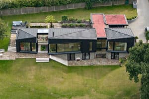 Aerial view of a modern, angular house with black exterior, large windows, tiered stone patio, rooftop garden, and surrounding green lawn.