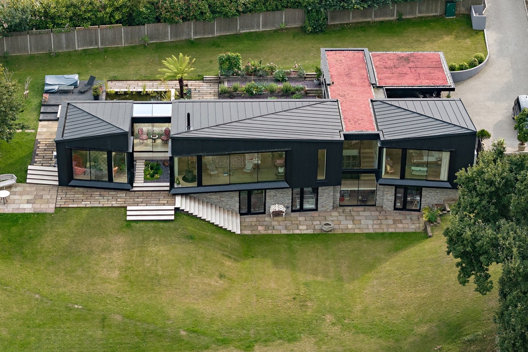Aerial view of a modern, angular house with black exterior, large windows, tiered stone patio, rooftop garden, and surrounding green lawn.