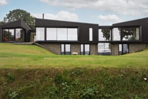 Modern house with large glass windows and dark exterior, situated on a grassy hill with stone foundation and steps leading to the entrance.