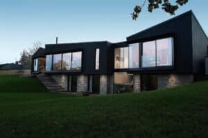 Modern two-story house with large glass windows, dark siding, and stone accents, situated on a grassy slope at dusk. Indoor lights illuminate the interior.