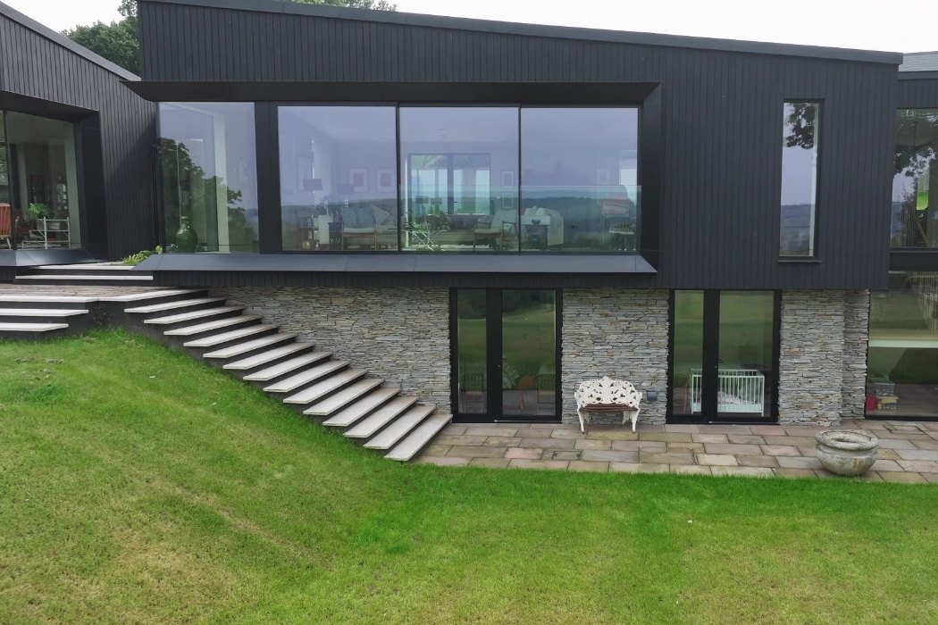 Modern two-story house with large glass windows, black siding, stone facade on lower level, outdoor staircase, and green lawn.