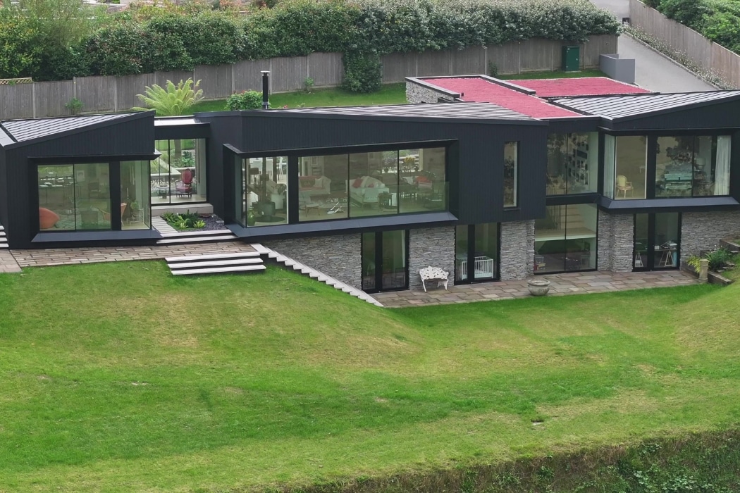 Modern house with large glass windows, dark exterior panels, stone accents, and a flat roof, set on a sloped grassy lawn with landscaped garden areas.