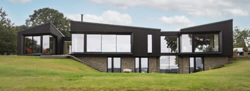 A modern two-story house with dark exterior panels, large glass windows, and stone accents, set on a grassy lawn with an overcast sky.