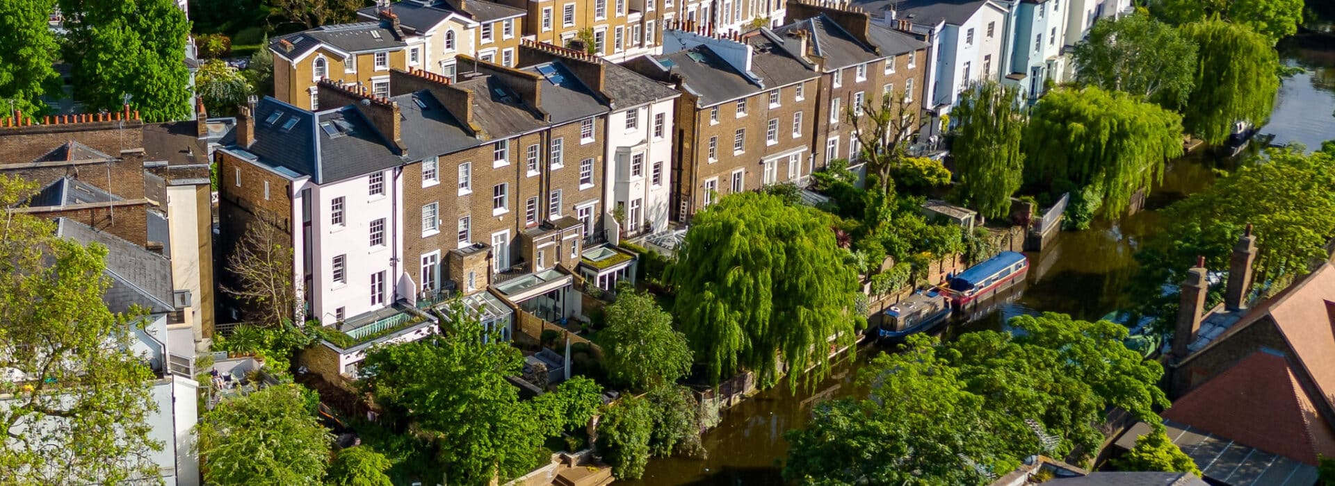 A row of townhouses with gardens lines a canal, where a narrowboat is moored among trees and greenery.