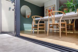 Open glass doors reveal a modern dining area with wooden chairs, a white table, artwork on the walls, and a large circular mirror reflecting the interior.