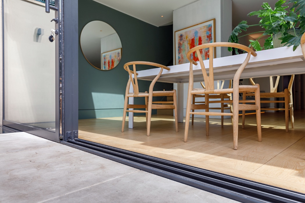 Open glass doors reveal a modern dining area with wooden chairs, a white table, artwork on the walls, and a large circular mirror reflecting the interior.