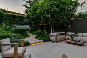 A modern outdoor patio with cushioned seating, surrounded by lush greenery and illuminated steps; two dogs rest on the sofa.