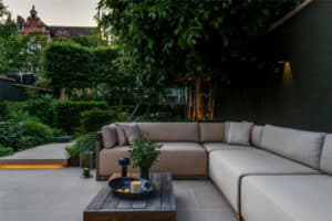 Modern outdoor patio with beige sectional sofas, a wooden coffee table holding a candle and plant, surrounded by lush greenery and trees in a landscaped garden setting.