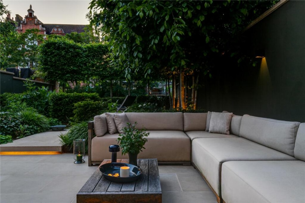 Modern outdoor patio with beige sectional sofas, a wooden coffee table holding a candle and plant, surrounded by lush greenery and trees in a landscaped garden setting.