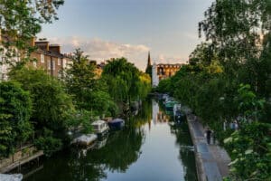 View of a canal lined with boats and trees, residential buildings on the left, a pedestrian path on the right, and a church spire in the background at sunset.