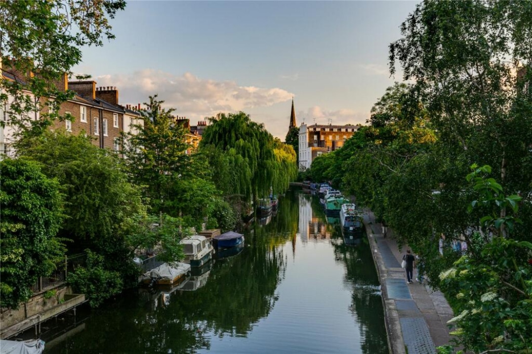 View of a canal lined with boats and trees, residential buildings on the left, a pedestrian path on the right, and a church spire in the background at sunset.