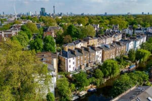Aerial view of a row of townhouses along a canal with trees and greenery, set against a city skyline in the background.