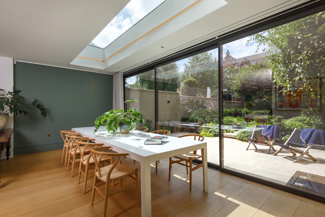Modern dining room with a long white table, wooden chairs, indoor plants, large skylight, and floor-to-ceiling glass doors opening to a sunny garden patio.