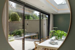 View through a round mirror of a modern dining area with a white table, green plants, and sliding glass doors opening to a patio garden.