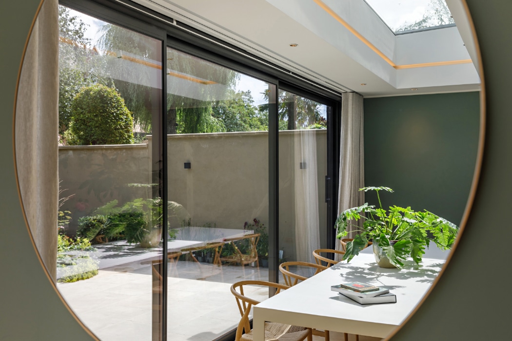 View through a round mirror of a modern dining area with a white table, green plants, and sliding glass doors opening to a patio garden.