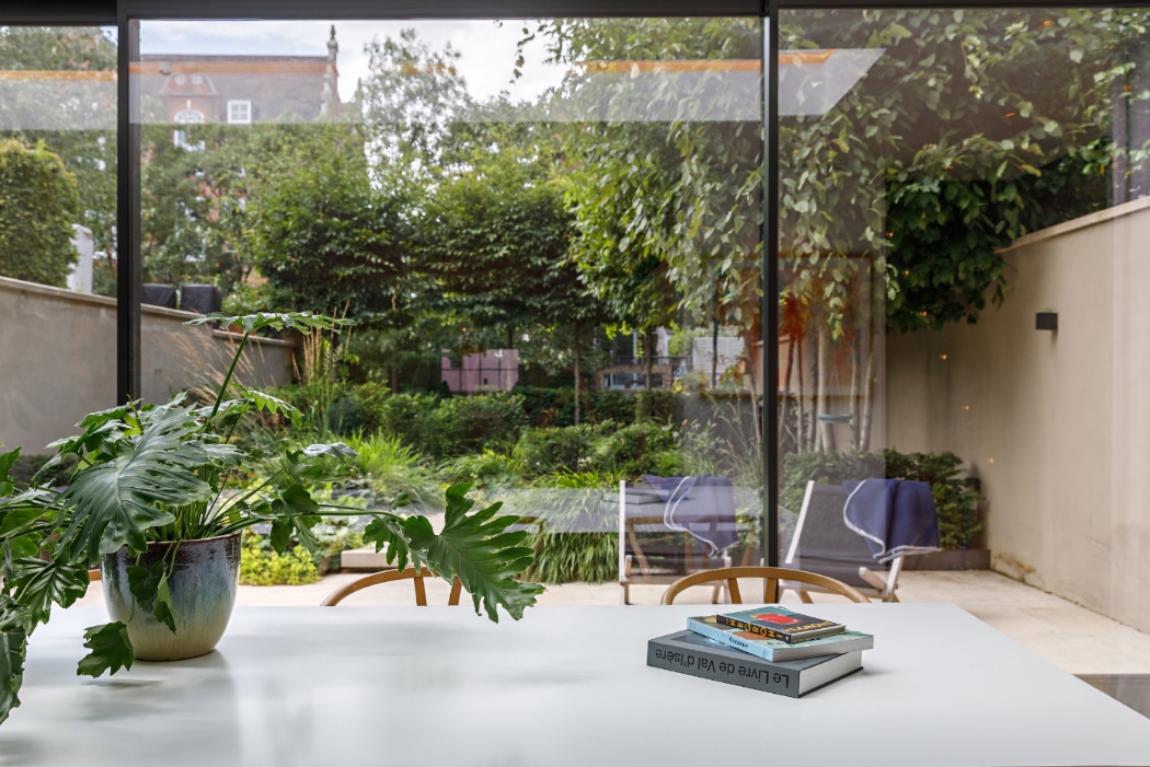 A white table with a potted green plant and stacked books sits by floor-to-ceiling glass doors, overlooking a leafy garden with outdoor chairs.