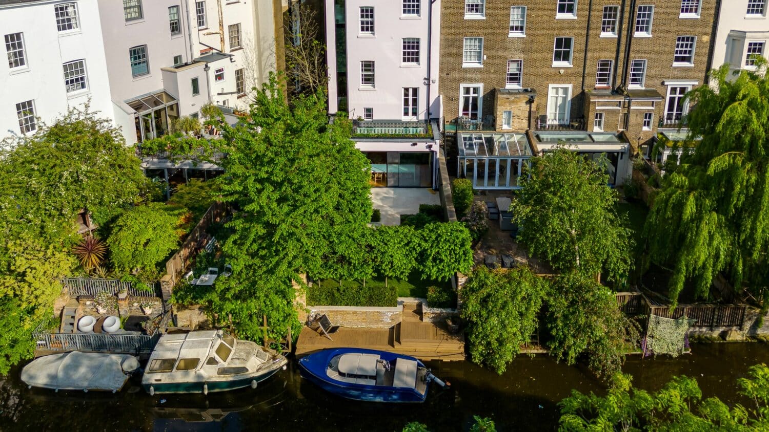 A riverside view of residential buildings with gardens, patios, and two boats docked along the water.