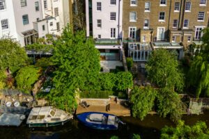 A riverside view of residential buildings with gardens, patios, and two boats docked along the water.
