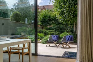 Modern dining area with a wooden table and chair next to a glass door opening to a patio with two lounge chairs and towels, overlooking a lush green garden.
