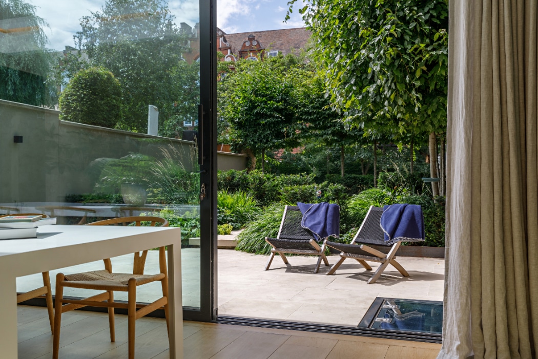 Modern dining area with a wooden table and chair next to a glass door opening to a patio with two lounge chairs and towels, overlooking a lush green garden.