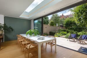 Modern dining area with a long white table, wooden chairs, houseplants, and large glass doors opening to a lush garden patio with lounge chairs. Skylight lets in natural light.