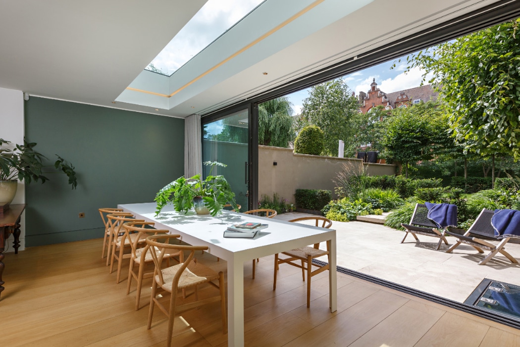 Modern dining area with a long white table, wooden chairs, houseplants, and large glass doors opening to a lush garden patio with lounge chairs. Skylight lets in natural light.