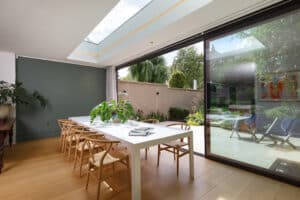 Modern dining area with a long white table and wooden chairs, large skylight, and glass doors opening to a sunny garden patio.
