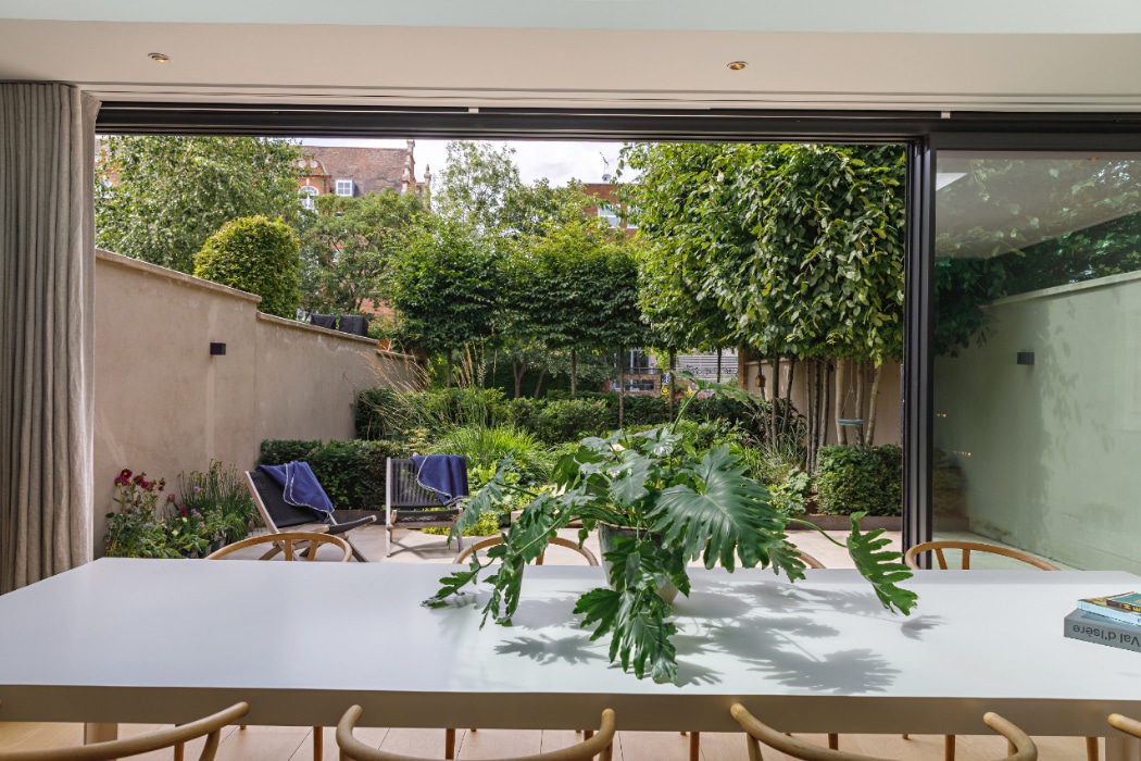 A modern dining area with a white table and green plant centerpiece overlooks a well-maintained garden through large glass sliding doors.