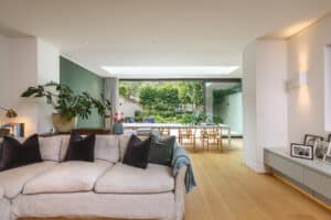 Modern living room with a beige sofa, green accent wall, and large glass doors opening to a dining area and garden. Wooden floors and indoor plants are visible.