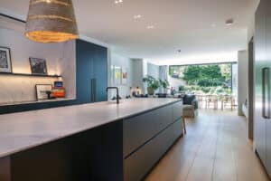 Modern open-plan kitchen with a large marble island, dark cabinets, light wood flooring, and a view of a living area and garden through sliding glass doors.