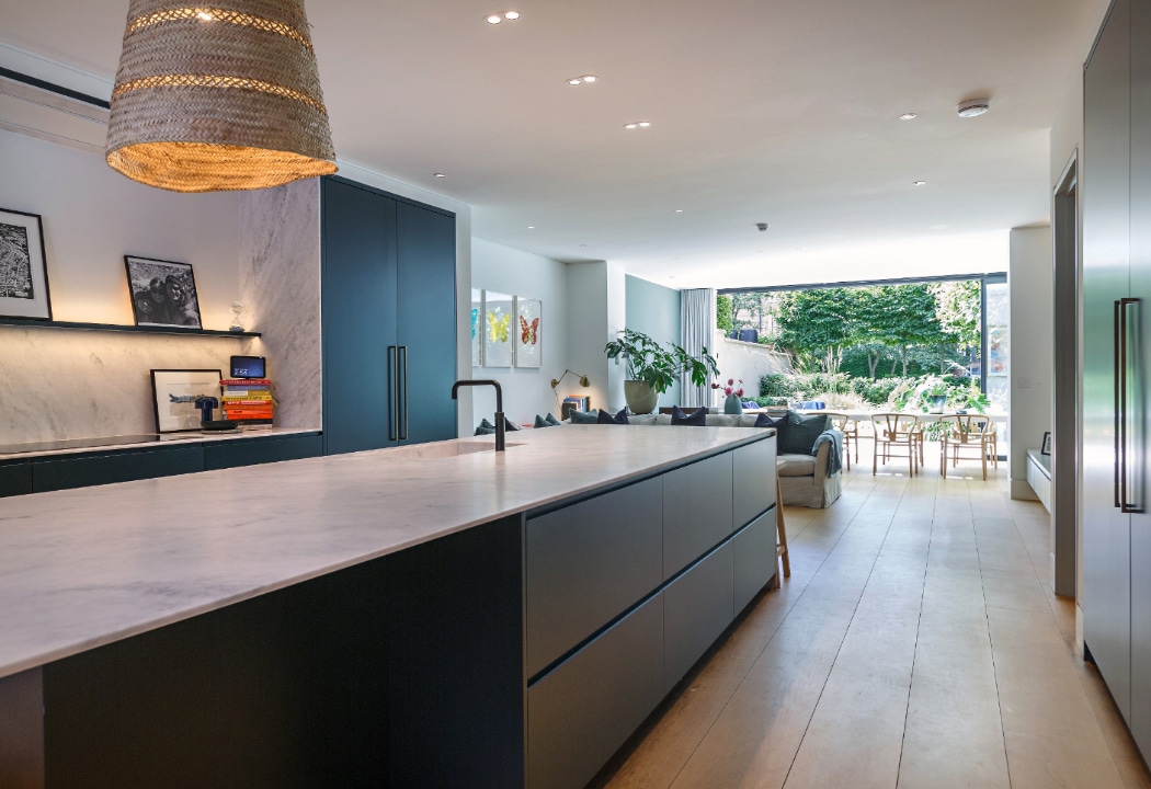 Modern open-plan kitchen with a large marble island, dark cabinets, light wood flooring, and a view of a living area and garden through sliding glass doors.
