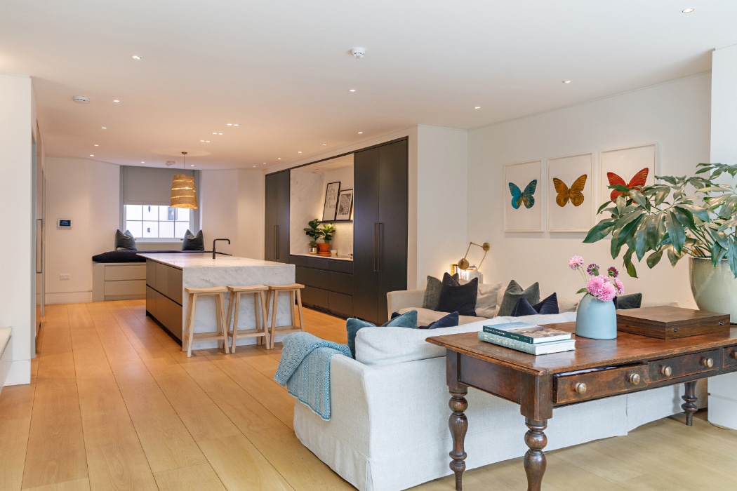 Open-plan living area with wooden floors, modern kitchen with island, beige sofa, wooden desk, potted plant, and butterfly art on the wall.