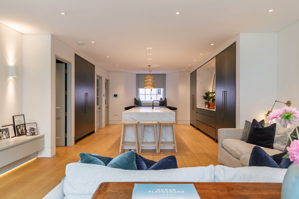 Modern open-plan living room and kitchen with light wood floors, neutral furnishings, built-in cabinets, a central island, and pendant lighting. Framed photos and flowers add decorative touches.
