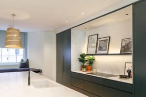 Modern kitchen with dark cabinets, white marble countertops and backsplash, potted plants, framed photos, and a woven pendant light fixture above a central island.
