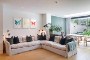A modern living room with a large light grey corner sofa, black and green cushions, butterfly art on the wall, plants, and a view of the garden through glass doors.