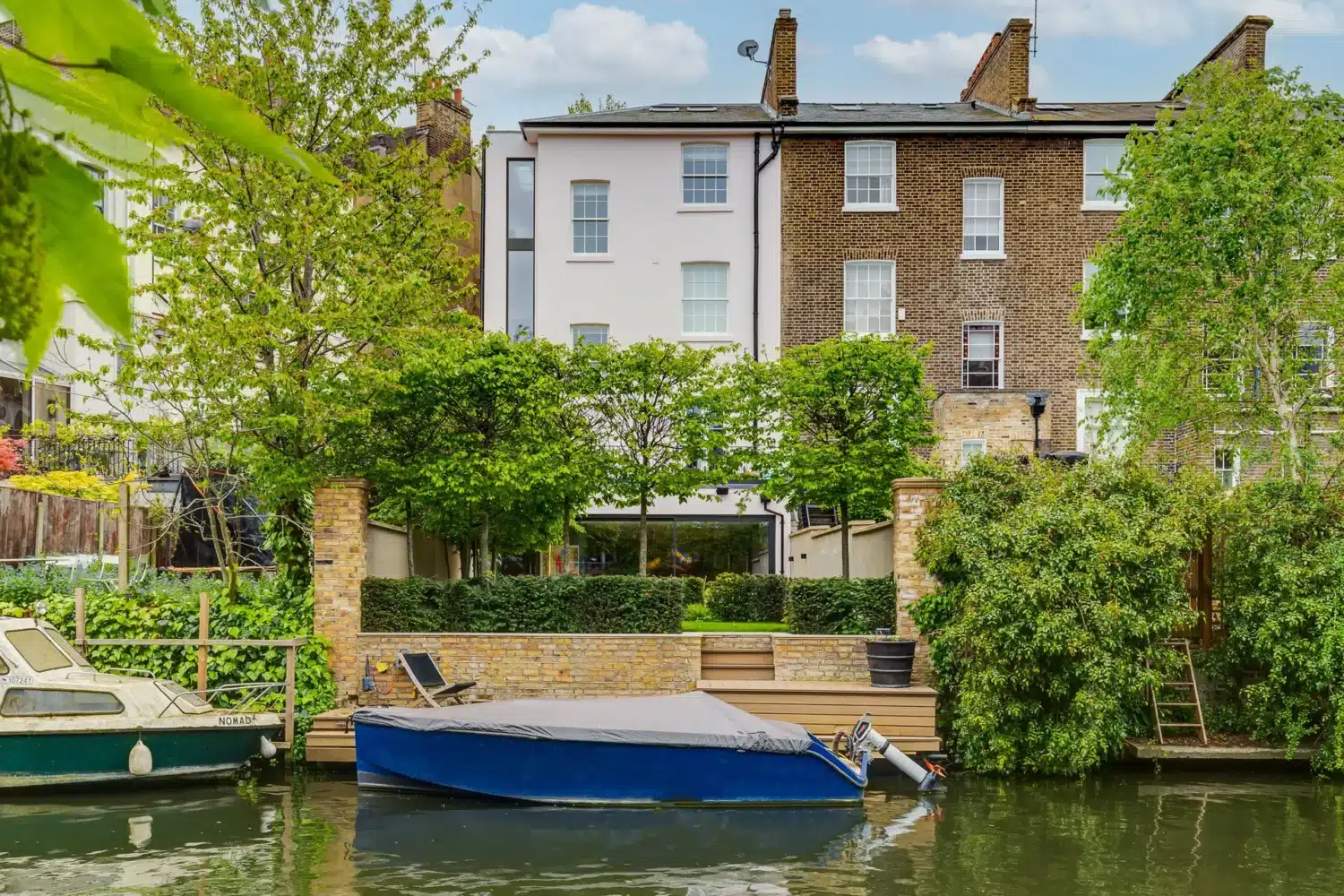 A riverside townhouse with a brick facade and large windows is seen behind a small dock with a covered boat and greenery along the water’s edge.