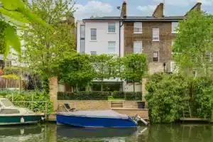 A riverside townhouse with a brick facade and large windows is seen behind a small dock with a covered boat and greenery along the water’s edge.