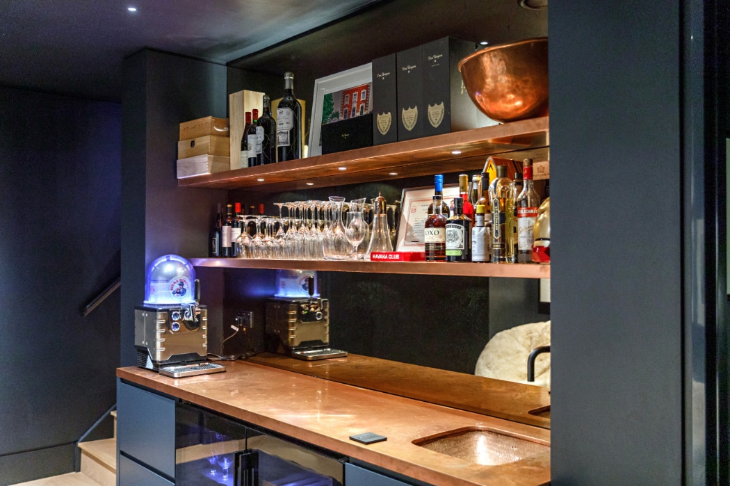 Modern home bar with a copper countertop, glassware, bottles of wine and spirits on shelves, two coffee machines, and a large copper bowl on the top shelf.