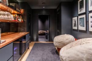 A modern lounge with a bar area, glassware, and bottles on shelves, two plush chairs, framed photos on the wall, and a view into an adjacent room with a large sofa.