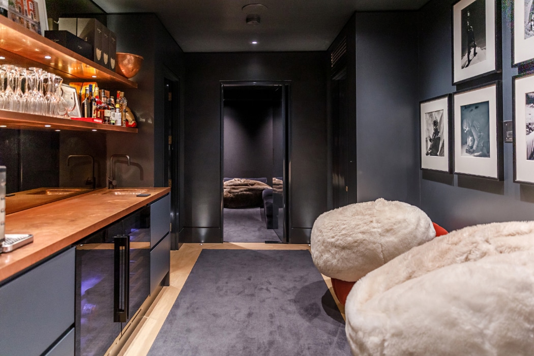 A modern lounge with a bar area, glassware, and bottles on shelves, two plush chairs, framed photos on the wall, and a view into an adjacent room with a large sofa.