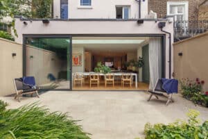 Modern house with large sliding glass doors open to a patio, revealing a dining area with a long table, chairs, plants, and artwork inside. Two lounge chairs sit outside.