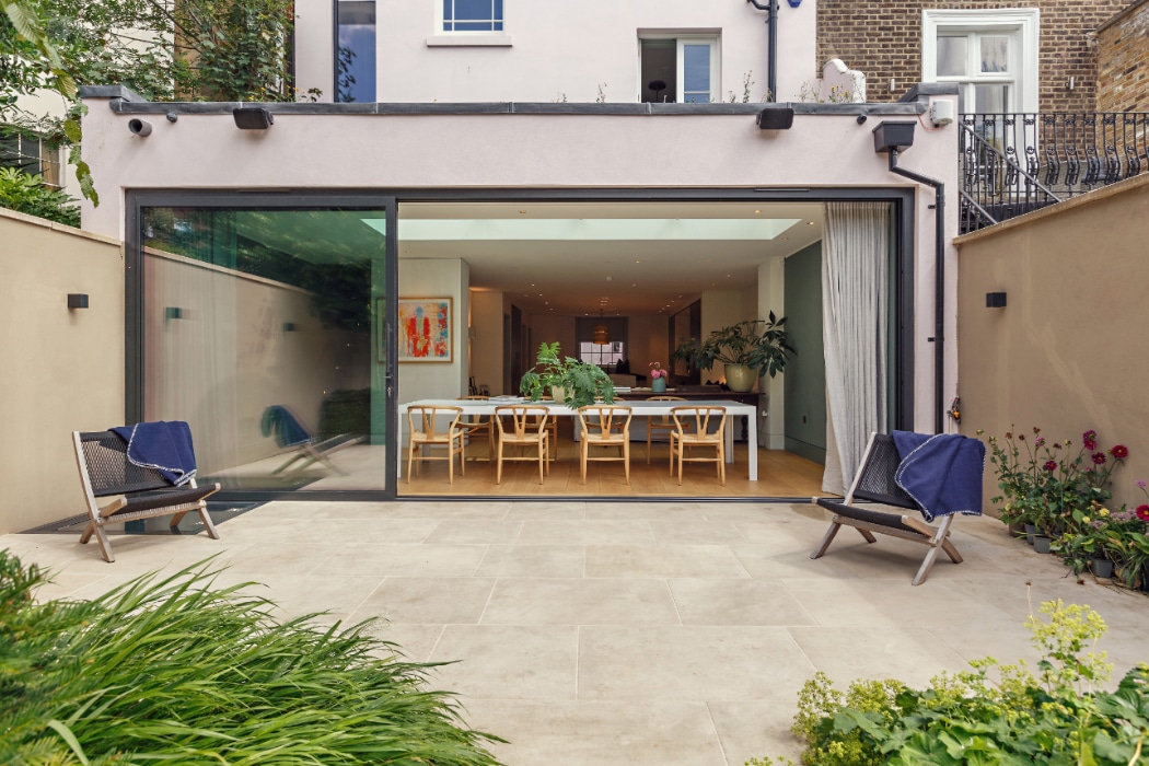 Modern house with large sliding glass doors open to a patio, revealing a dining area with a long table, chairs, plants, and artwork inside. Two lounge chairs sit outside.