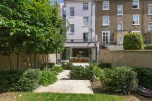 A modern townhouse with a landscaped garden, featuring a concrete path leading to a dining area inside glass doors at the back of the house.