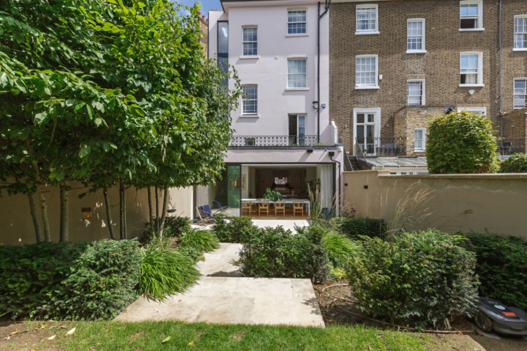 A modern townhouse with a landscaped garden, featuring a concrete path leading to a dining area inside glass doors at the back of the house.