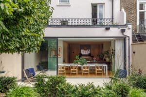 Modern home with a large glass sliding door opening to a patio and garden, featuring a dining table with wooden chairs and indoor plants.