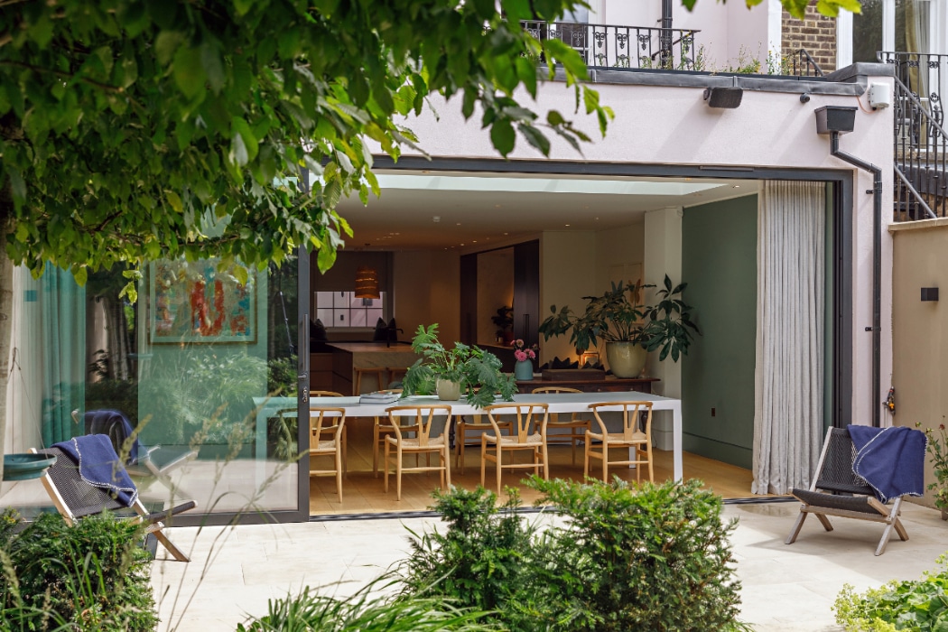 A modern open-plan dining area with a long table and wooden chairs opens onto a garden patio through large sliding glass doors. Plants decorate the space indoors and outdoors.