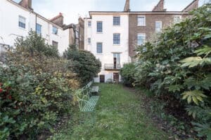 A garden with green benches and overgrown bushes leads to the back entrance of a multi-story brick and white townhouse.