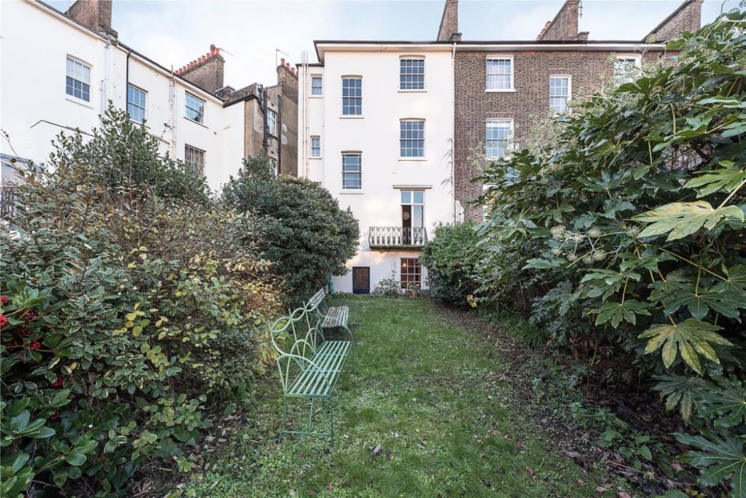 A garden with green benches and overgrown bushes leads to the back entrance of a multi-story brick and white townhouse.