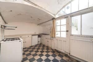 A worn kitchen with checkered floor, white cabinets, an old stove, and large windows letting in natural light; walls and ceiling show visible signs of wear and peeling paint.