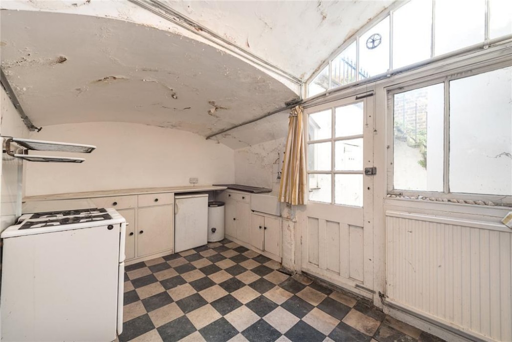 A worn kitchen with checkered floor, white cabinets, an old stove, and large windows letting in natural light; walls and ceiling show visible signs of wear and peeling paint.